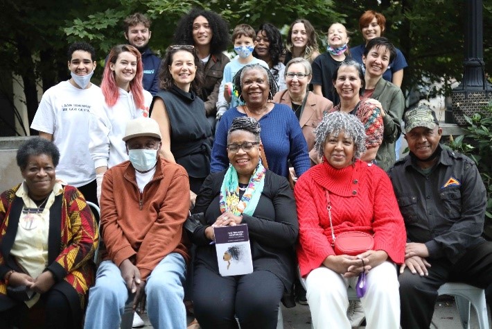 A group of people, young and older adults, seated and standing outside in front of foliage.